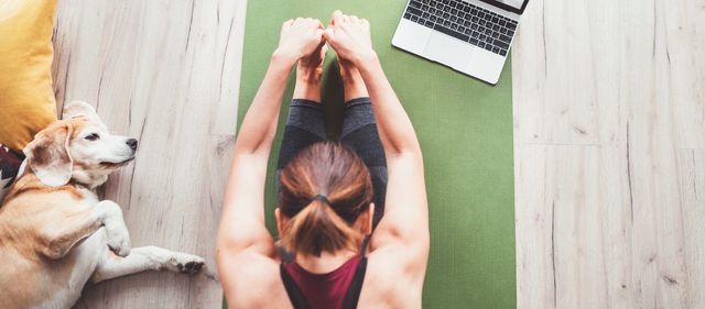 Una mujer está haciendo yoga en una colchoneta junto a un perro y una computadora portátil.